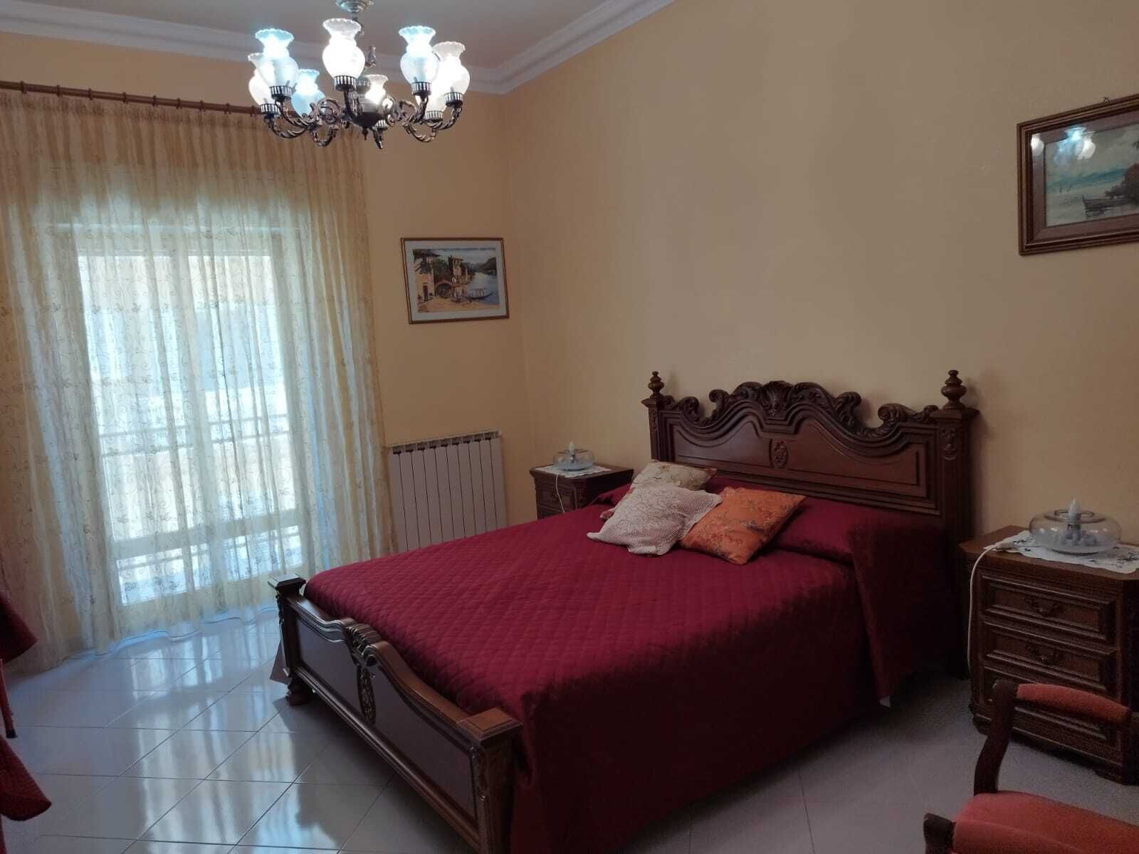 Une chambre à coucher avec un grand lit, un lustre, un radiateur, une commode en bois et une fenêtre avec des rideaux.