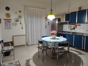 A dining area with a round table, chairs, a flower vase, and a kitchen with blue cabinets and a hanging light fixture.