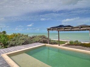 Una piscina infinita con terraza de madera y un pérgola con sombrillas, con vistas al océano.