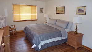 Un dormitorio con cama, mesitas de noche, cómoda de madera, ventana con persianas y un cuadro colgado en la pared.