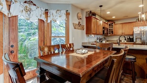 A wooden dining table with six chairs, a kitchen with wooden cabinets, a refrigerator, and a window with a view of greenery.