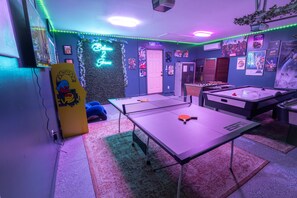 A room with a ping pong table, foosball table, and a neon sign on the wall.