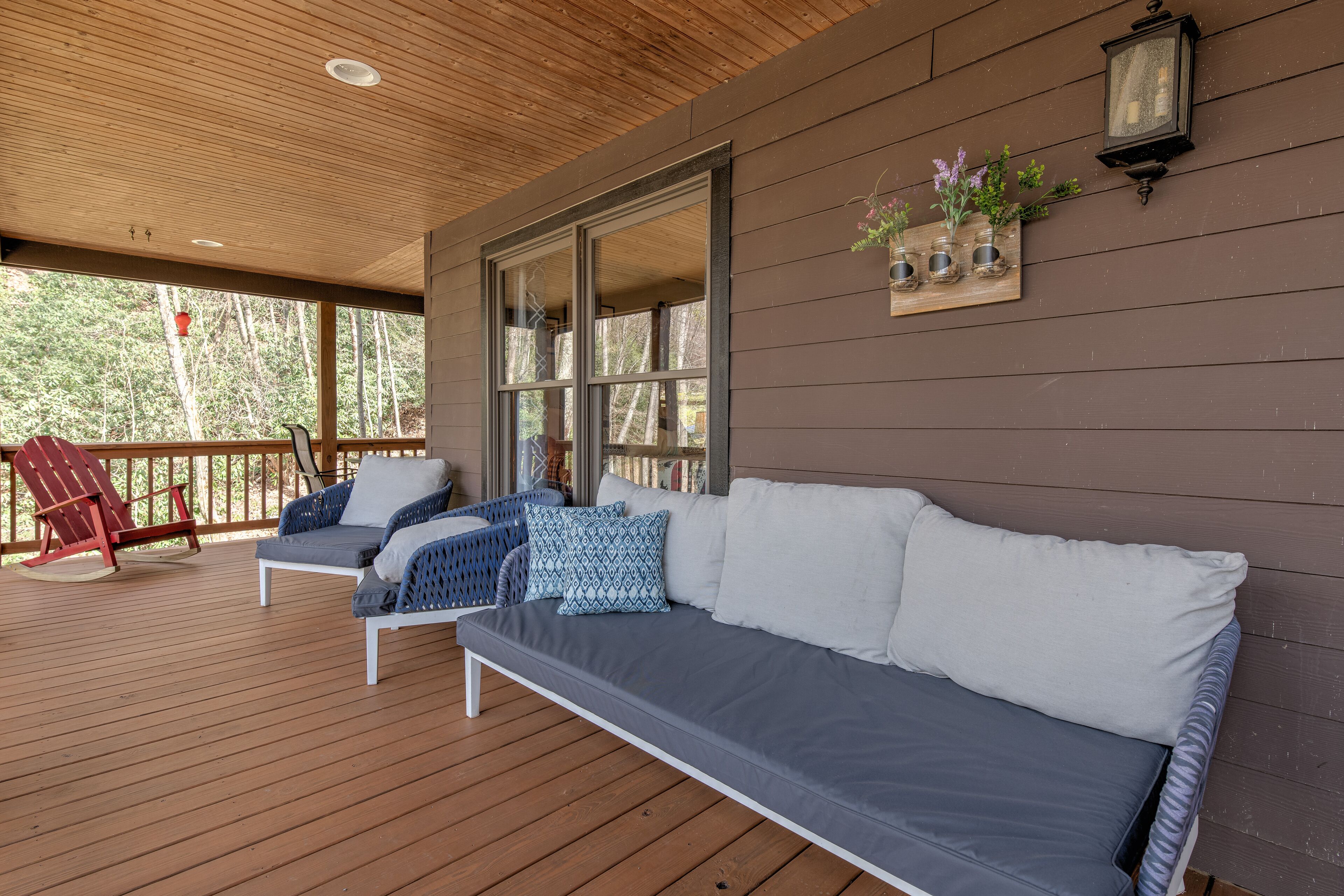 Un portico coperto con un pavimento in legno, un divano grigio con cuscini bianchi, una poltrona Adirondack rossa e una fioriera a parete con fiori.