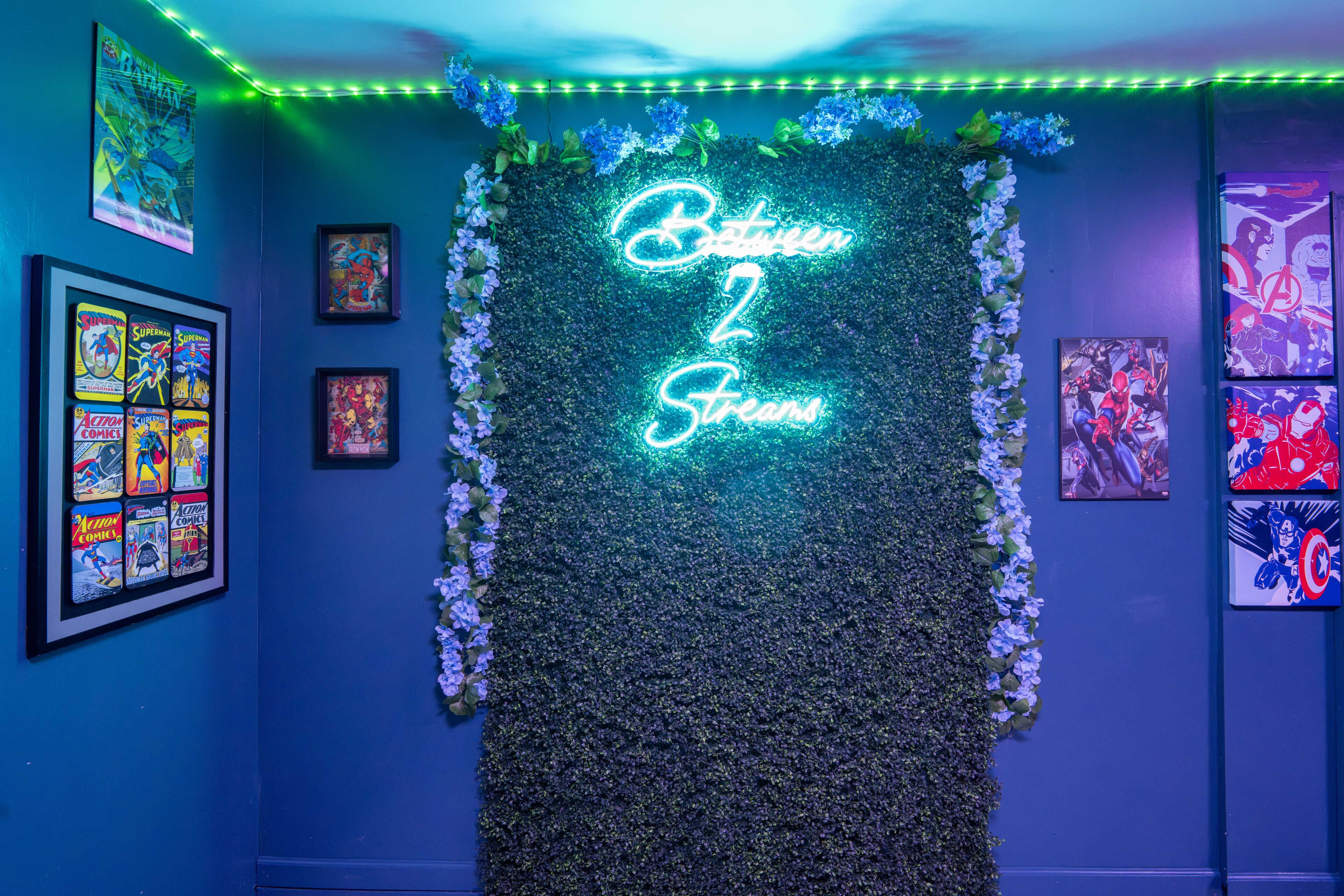 A room with a neon sign reading 'Between 2 Straws,' framed comic book posters, and a wall decorated with white flowers.