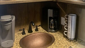 Eine Küche mit einer Granitarbeitsplatte, einem Kupferspülbecken, einer Kaffeemaschine, einer Kaffeetasse, einem Willkommensschild und diversen Küchenutensilien auf Holzregalen.