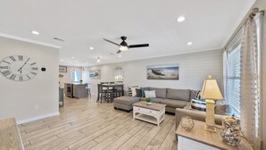 A spacious living room with a wooden floor, a ceiling fan, a large clock, and a dining area in the background.