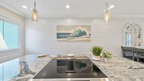 A kitchen with a built-in stove, a painting of a wave, pendant lights, and a window with blinds.