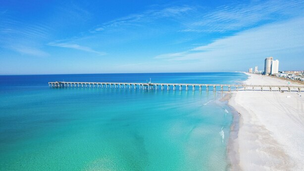 A coastal scene with a long pier extending into the ocean, a sandy beach, and a cityscape with buildings in the background.