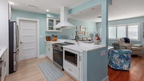 A modern kitchen with white cabinets, a built-in oven, and a microwave. There is a seating area with a blue patterned ottoman and a beige sofa.