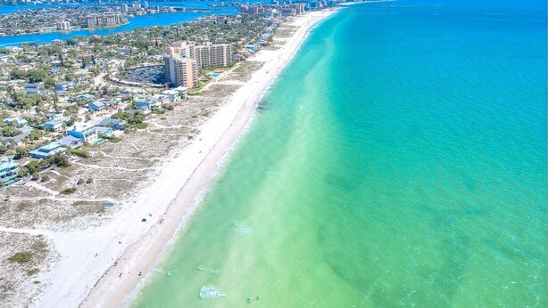 An aerial view of a coastal city with a long sandy beach, clear blue waters, and a dense urban area with high-rise buildings.