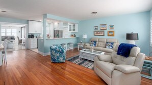 A spacious living room with a beige sofa, a coffee table, and a blue patterned ottoman. There is a kitchen area with white cabinets and a dining area with a table and chairs.