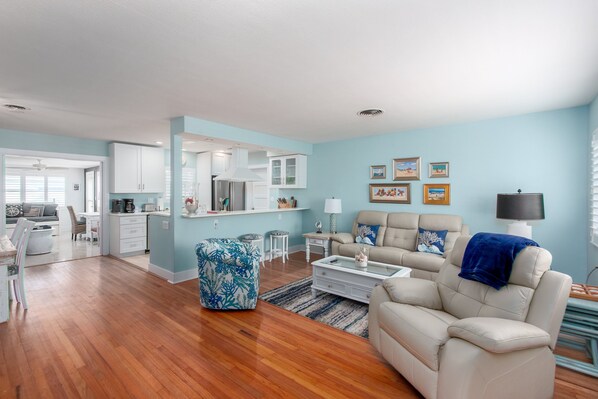 A spacious living room with a beige sofa, a coffee table, and a blue patterned ottoman. There is a kitchen area with white cabinets and a dining area with a table and chairs.