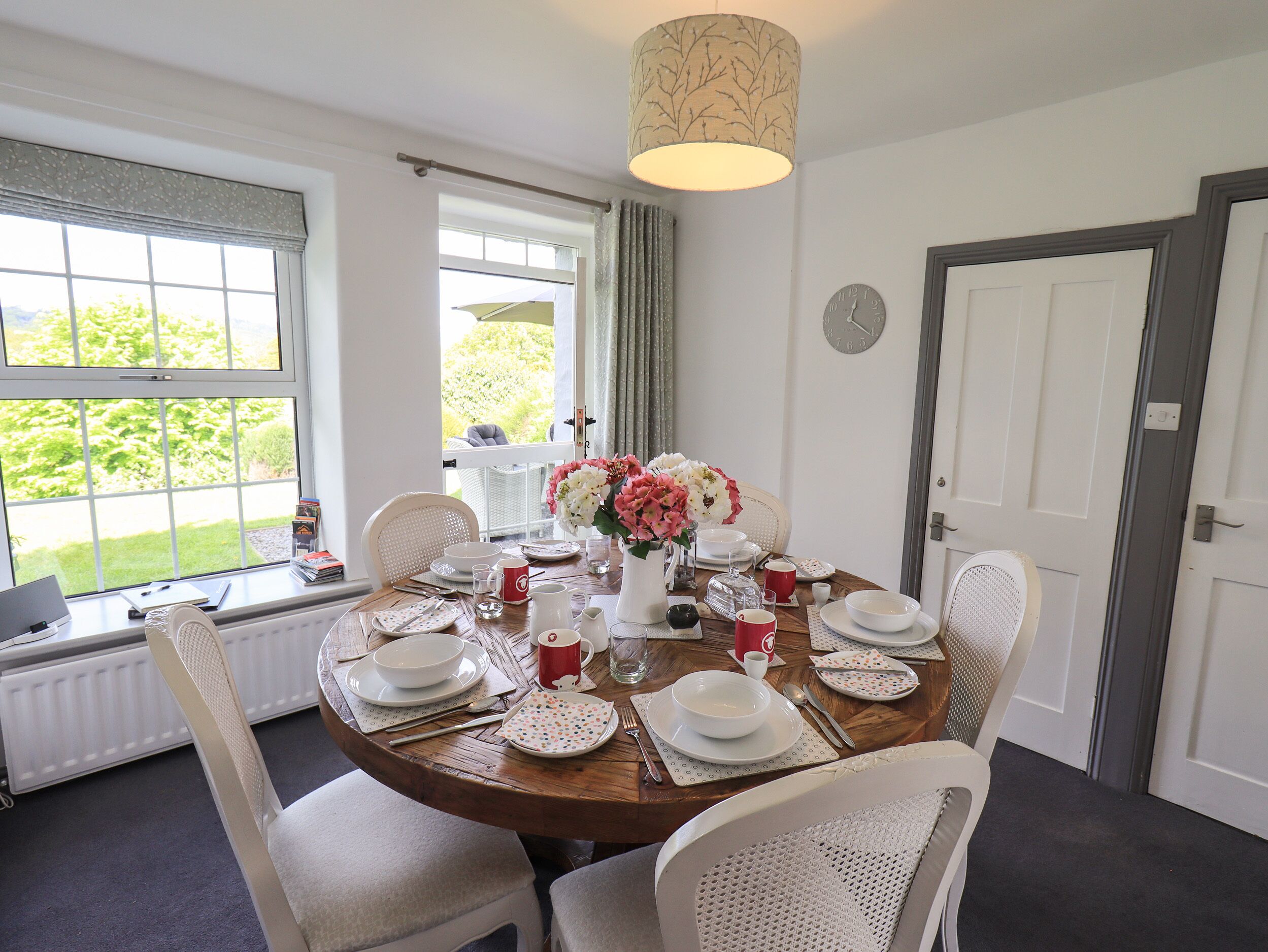 A dining room with a round table set for four, white chairs, a vase with flowers, and a clock on the wall.