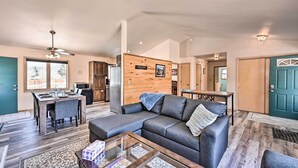 Un grand espace de vie comprenant un canapé, une table basse et une table à manger. Un mur en bois, orné d’un tableau, se trouve à droite, ainsi qu’une porte.