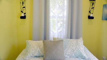 A bed with patterned bedding, a window with sheer curtains, and wall-mounted light fixtures.