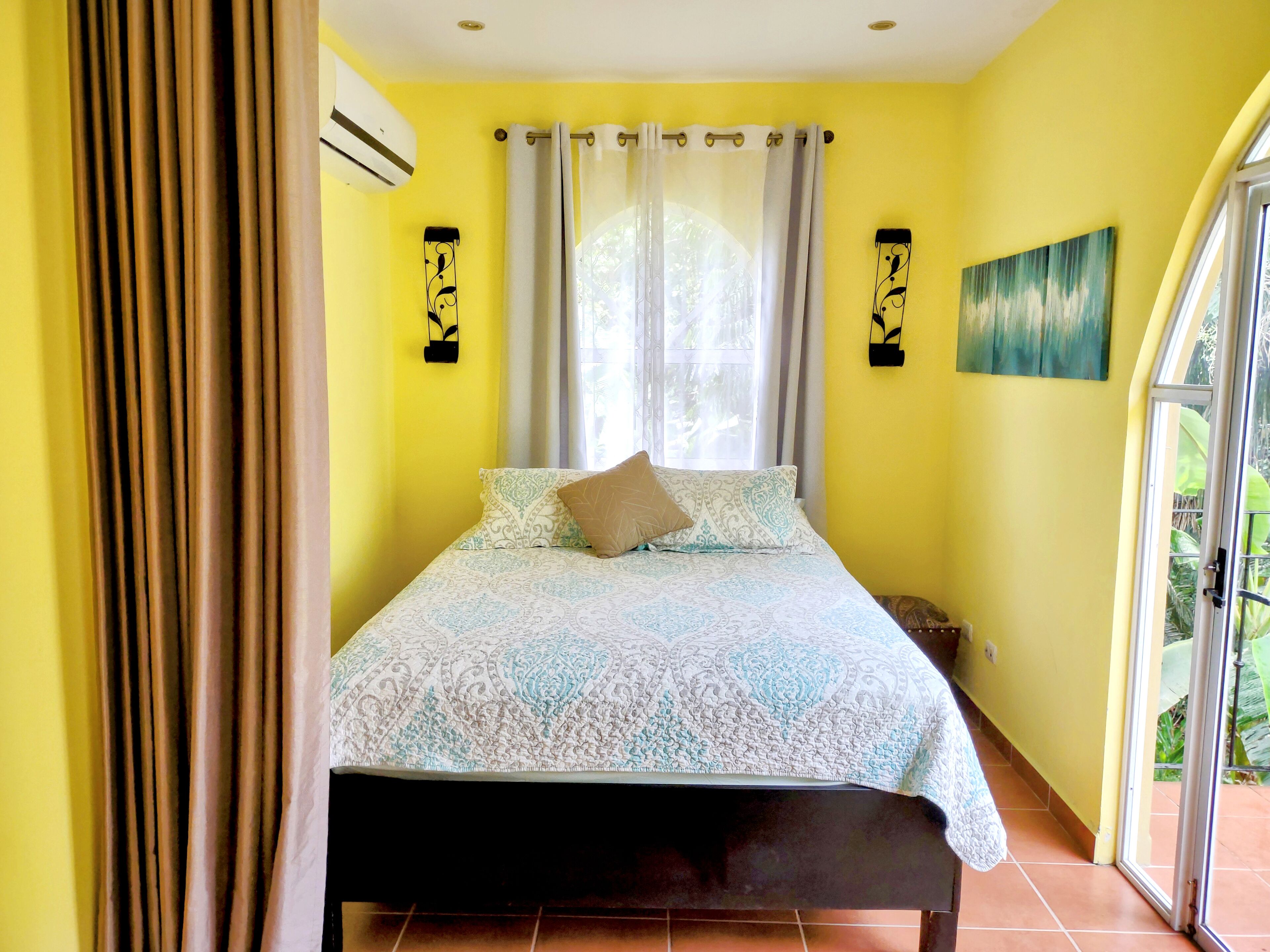 Un dormitorio con cama, cortinas, un equipo de aire acondicionado y un cuadro en la pared.
