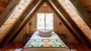 A bedroom with a sloped ceiling, a window, a bed with a patterned blanket, and two bedside tables. - Come enjoy nature in our cabin !  (Idyllwild-Pine Cove)