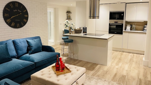 A modern living room with a blue sofa, a large wall clock, and a kitchen area with white cabinets and stainless steel appliances. - Escape London in London - 2 bed luxury apartment in lovely Wanstead (London)