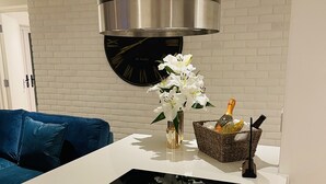 A modern kitchen with a black induction cooktop, a stainless steel range hood, a white brick wall, a blue sofa, and a vase with white flowers. - Escape London in London - 2 bed luxury apartment in lovely Wanstead (London)