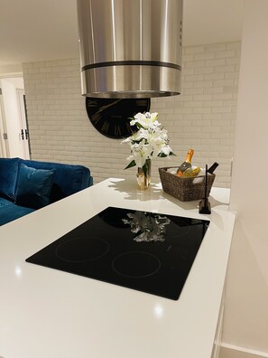 A modern kitchen with a black induction cooktop, a stainless steel range hood, a white brick wall, a blue sofa, and a vase with white flowers.