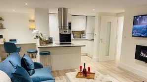 A modern living room with a blue sofa, a small ottoman with a tray of red wine glasses, and a kitchen in the background. - Escape London in London - 2 bed luxury apartment in lovely Wanstead (London)