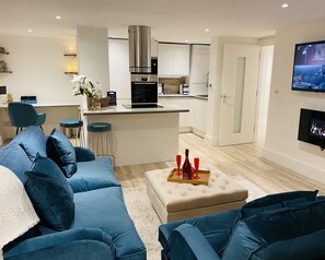 A modern living room with a blue sofa, a small ottoman with a tray of red wine glasses, and a kitchen in the background.