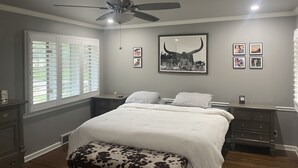 A bedroom with a bed, a ceiling fan, a window with blinds, a bedside table, a nightstand, and a wall with framed pictures. - Midtown Rancher with Great Backyard and Deck for Entertaining (Tulsa)