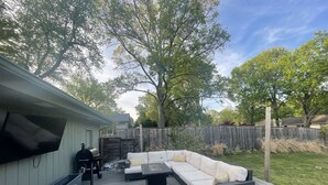 A backyard with a white outdoor sofa, a grill, and a TV mounted on the wall. - Midtown Rancher with Great Backyard and Deck for Entertaining (Tulsa)