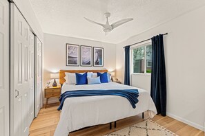 A bedroom with a bed, bedside tables, a ceiling fan, a window with curtains, and framed pictures on the wall.