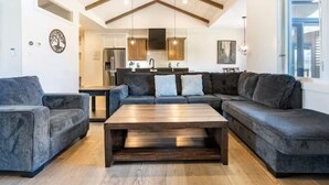 A modern living room with a large sectional sofa, a wooden coffee table, and a kitchen in the background. - Laporte Cottage | Hot Tub | Walk to Beach | Fire Pit | Grand Bend & Bayfield (Zurich)