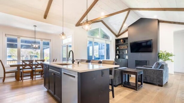A modern kitchen with a central island, wooden dining table, and chairs, a built-in TV, and a fireplace. - Laporte Cottage | Hot Tub | Walk to Beach | Fire Pit | Grand Bend & Bayfield (Zurich)