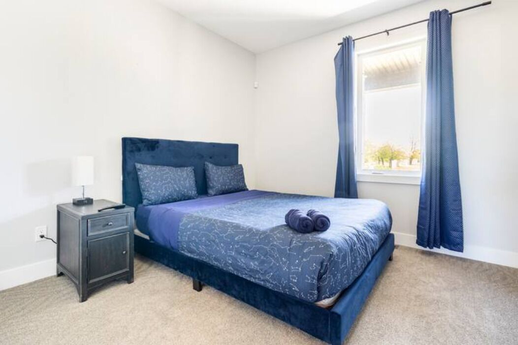 Un dormitorio con cama, mesita de noche, ventana con cortinas y vista a árboles.