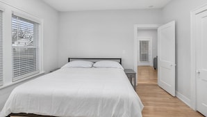 Ein Schlafzimmer mit einem Bett, einem Nachttisch, einem Fenster mit Jalousien und einer Tür zu einem anderen Raum.