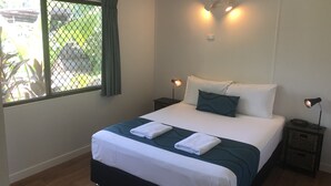A neatly made bed with white and teal bedding, two bedside lamps, and a window with a view of greenery. - Cocos Holiday Apartments (Trinity Beach)