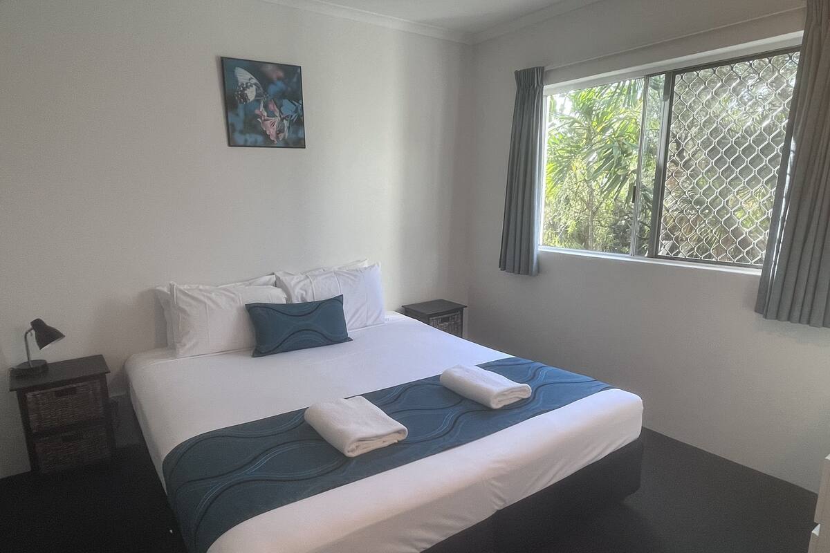 A neatly made bed with white and blue bedding, a small bedside table with a lamp, and a window with a view of greenery.
