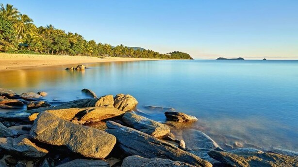 A beach with clear water, rocks, and palm trees. - Cocos Holiday Apartments (Trinity Beach)