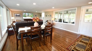 A spacious living room with a dining area, hardwood floors, and large windows offering a view of the outdoors. - Kayaks - Firepit - Dock - Fishing & more! (Lindstrom)