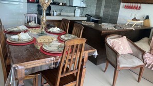 A dining area with a wooden table set for a meal, surrounded by chairs. A kitchen with dark wooden cabinets and a tiled backsplash is visible in the background. - Desfrute dessa maravilha. (Angra dos Reis)