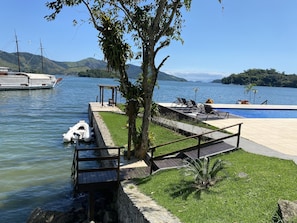 A lakeside pool area with a wooden deck, lounge chairs, and a small boat docked near the water. - Desfrute dessa maravilha. (Angra dos Reis)