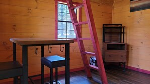 Une pièce aux murs en bois, avec une fenêtre, une table, un tabouret, une échelle et une étagère.