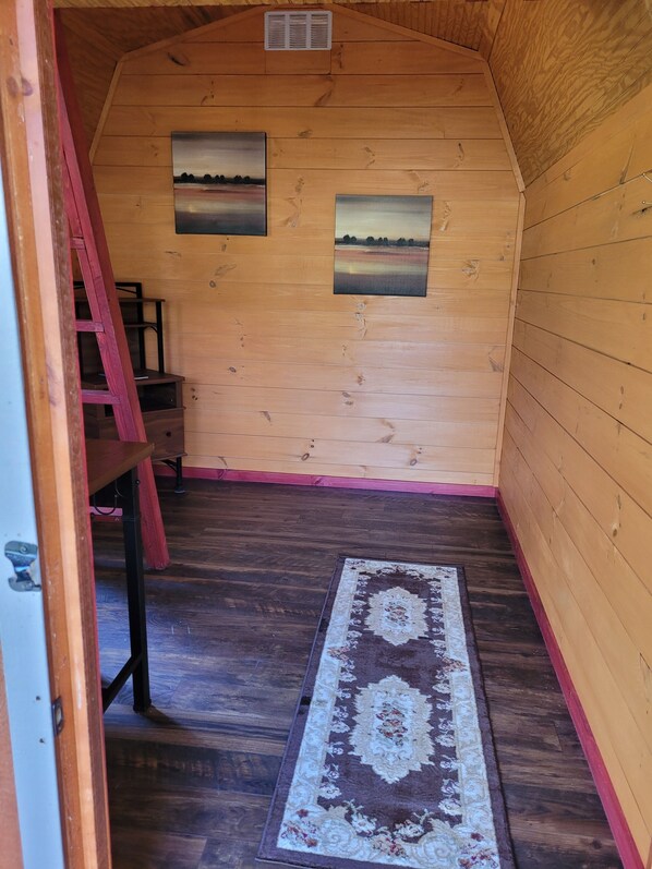 Un interior de madera con suelo de madera, una escalera roja, dos cuadros en la pared y una alfombra estampada.