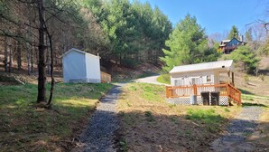 Un chemin de gravier mène à un chalet doté d’une terrasse en bois et d’une remise, le tout situé dans une zone boisée.