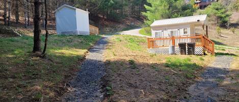 Un chemin de gravier mène à un chalet doté d’une terrasse en bois et d’une remise, le tout situé dans une zone boisée.