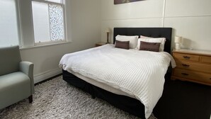 A bedroom with a large bed, a nightstand, a chair, a window with stained glass, and a painting of a forest scene. - Bayview house in Oriental Parade  (Wellington)