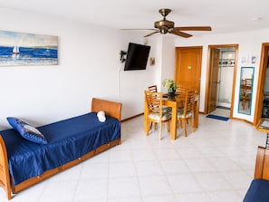 A room with a bed, a dining table with chairs, a ceiling fan, a TV mounted on the wall, and a painting of a sailboat.