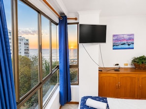 A room with a bed, a TV mounted on the wall, a painting, and a view of the ocean.