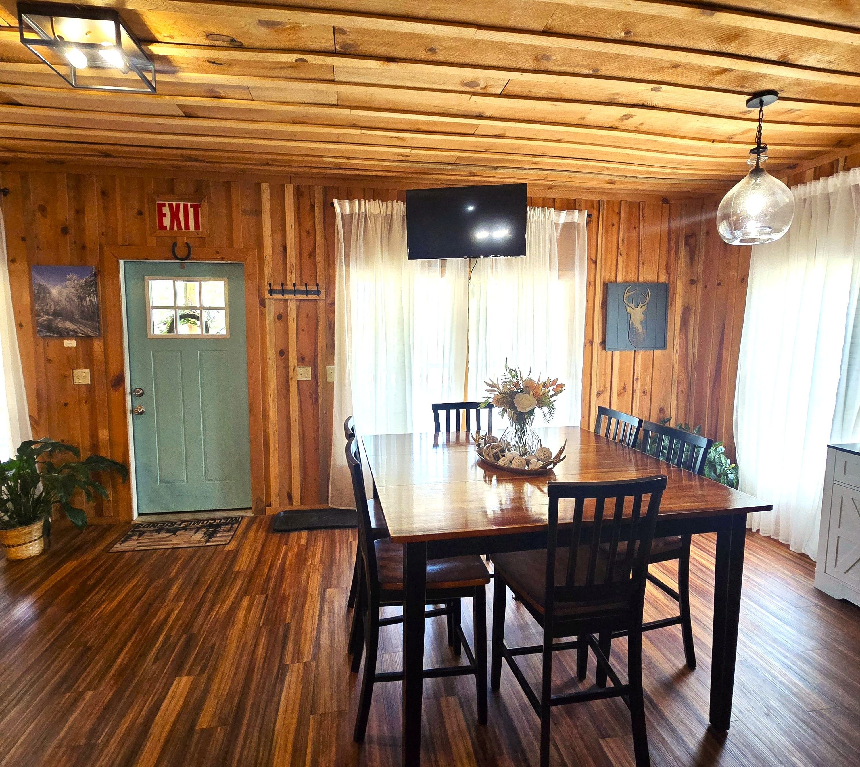 A wooden-paneled room with a dining table, chairs, a TV, and a door with an exit sign.