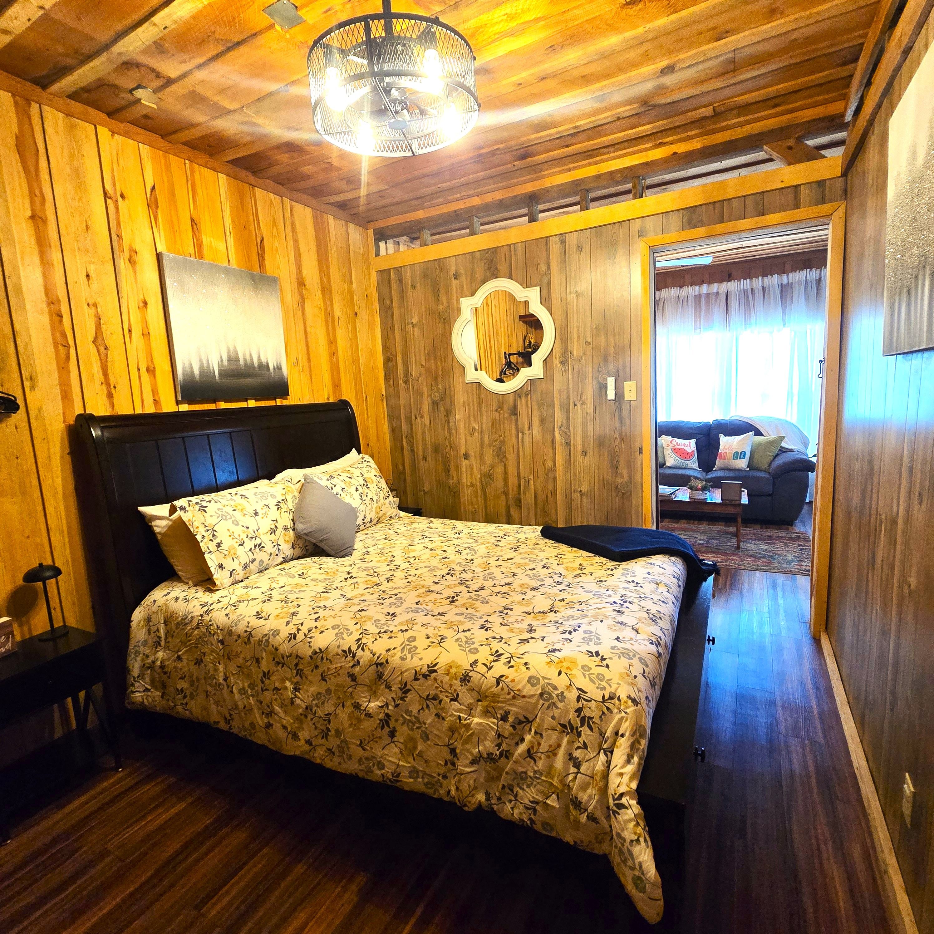 A wooden-paneled bedroom with a bed, a wall mirror, and a view into another room.