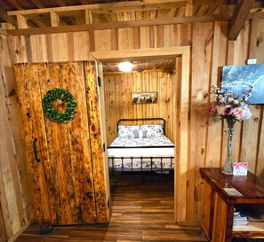 Una habitación de madera con una cama, una puerta de madera con un adorno de flores verdes, un cuadro en la pared y una mesita con un florero.