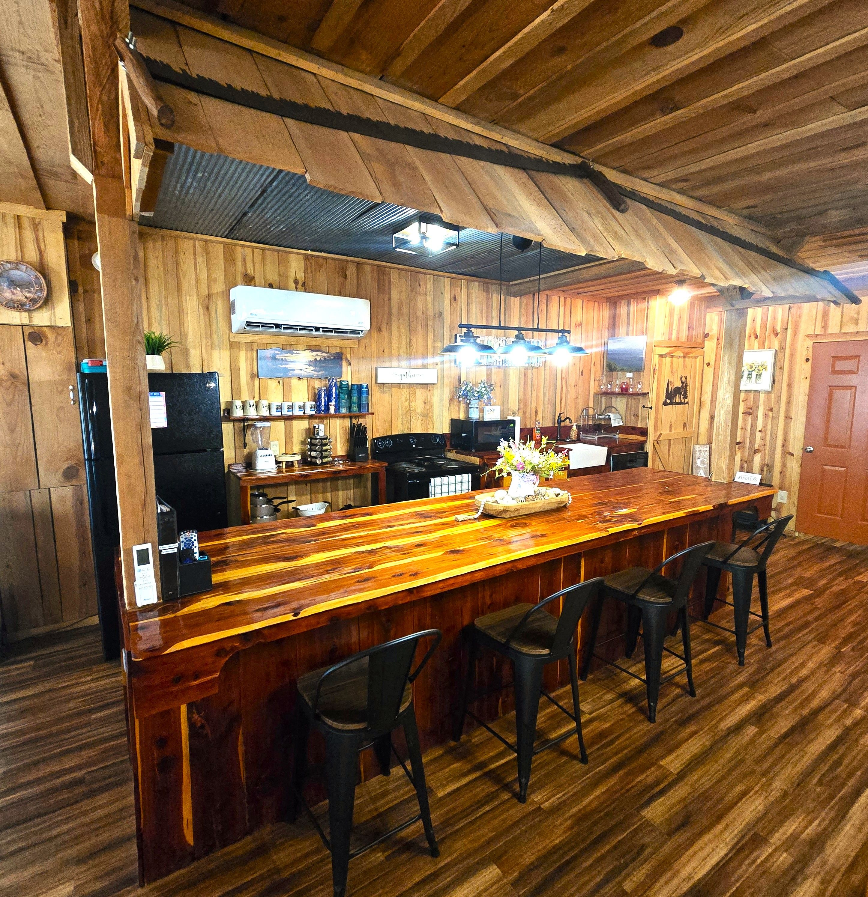 A wooden bar area with a long counter, bar stools, and a kitchen in the background.
